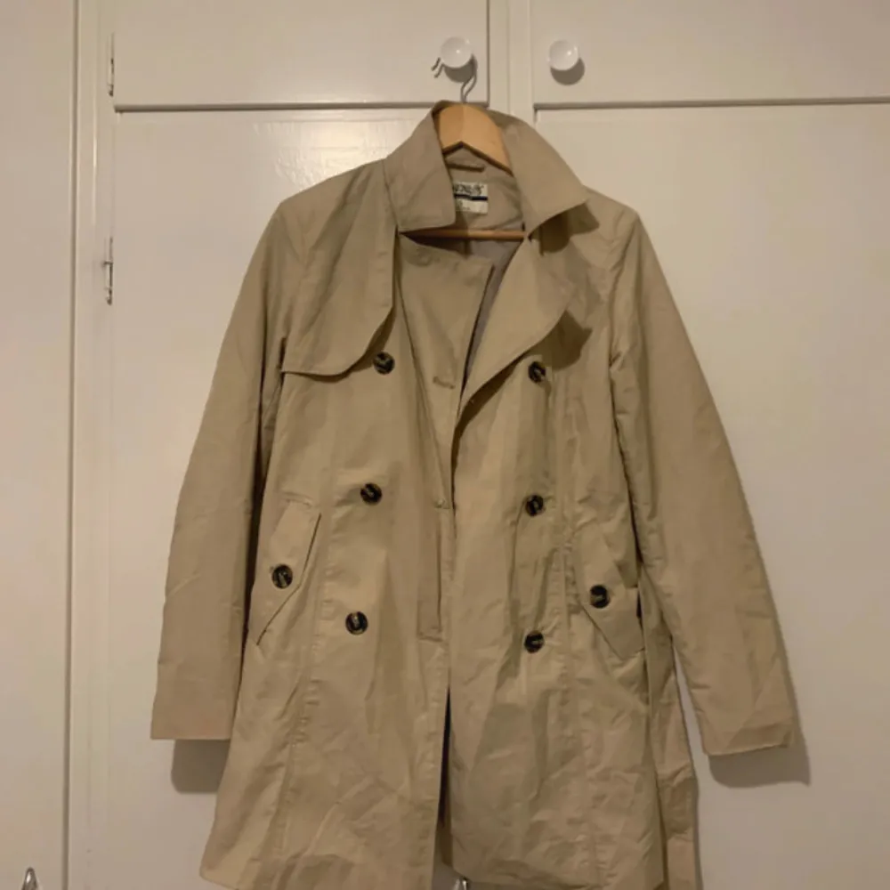 Säljer en klassisk beige trenchcoat i bra skick. Den har en dubbelknäppt design med stora knappar och två fickor fram. Perfekt för höst och vår, och ger en stilren look. Passar både till vardags och lite finare tillfällen.. Jackor.