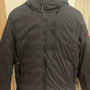 Svart dunjacka från Canada Goose - Säljer en svart dunjacka från Canada Goose i mycket bra skick. Jackan har en dragkedja framtill och en praktisk huva. Perfekt för kalla vinterdagar. Den har en klassisk design med en logotyp på ärmen. Passar både till vardags och mer äventyrliga aktiviteter.