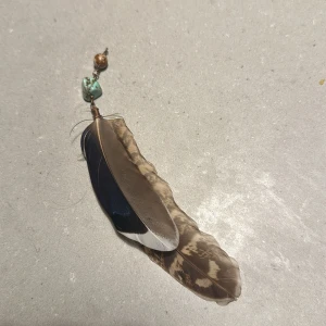 Örhänge med fjädrar och pärlor - Unikt örhänge med en kombination av naturliga fjädrar i bruna och blå nyanser. Smycket har också dekorativa pärlor i turkos och kopparfärg. Perfekt för en bohemisk stil eller som ett statement piece till din outfit. Krok fattas