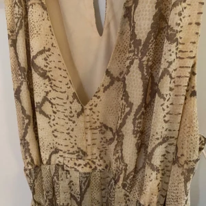 Ormmönstrad klänning från H&M - Snygg ormmönstrad klänning från H&M i beige och bruna toner. Den har en v-ringad design och ett knytband i midjan, denna är avtagbar. Perfekt för både vardag och fest! 