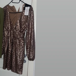  Nly trend paljettklänning - En superfin paljett klänning i färgen nougat/ljusbrun. Den har en djup v-ringning och långa ärmar, perfekt för festliga tillfällen som nyår! Klänningen är kort och har en tajt passform. Nyskick med prislapp kvar! ✨🤎