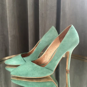 Gröna mockapumps med klack - Säljer ett par eleganta gröna mockapumps i bra skick, Stl 37. De har en spetsig tå och hög klack, perfekta för festliga tillfällen. Skorna är i en härlig grön nyans som verkligen sticker ut.