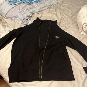 Svart zip up tröja från Prada - Säljer en stilren svart zip upp tröja från Prada. Den har en dragkedja framtill och ett snyggt logomärke på bröstet. Perfekt för både vardag och fest. Jackan är inte i så bra skick men passar till alla tillfällen. Storlek L men passar M