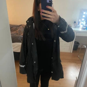 Svart regnjacka  - Säljer en svart regn jacka i bra skick. Den har en praktisk huva och långa ärmar med reflexdetaljer. Perfekt för höst och vinter, håller dig varm och torr. Jackan har knappar och dragkedja framtill för extra skydd mot väder och vind.
