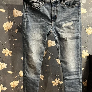 Blå jeans med slitningar - Säljer ett par snygga blå jeans i storlek EUR 152. De har coola slitningar på framsidan och en klassisk femficksdesign. Perfekta för en avslappnad stil och passar till både vardag och fest. Dragkedja och knapp framtill för enkel stängning.