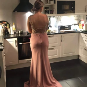 Rosa långklänning med pärlor - Säljer en fantastisk rosa långklänning med v-ringad design och pärldetaljer. Klänningen har en öppen rygg och är perfekt för festliga tillfällen. Materialet är mjukt och faller fint, vilket ger en elegant look. Perfekt för bal eller nyår!