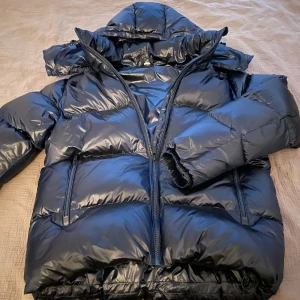 Marinblå pufferjacka med avtagbar huva - Säljer en snygg färgsvarierande (blå/svart) Everest pufferjacka i mycket bra skick. Den är perfekt för kalla vinterdagar och har en avtagbar huva. Jackan har en dragkedja framtill och är långärmad. Den är gjord av syntetmaterial som ger en glansig finish. Perfekt för både vardag och vinteräventyr!