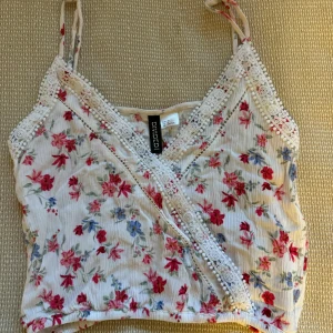 Blommig omlottopp från H&M Divided - Söt blommig omlottopp från H&M Divided i storlek 32. Toppen har ett vackert blommönster i rött och blått på en vit bakgrund. Den är ärmlös med spetsdetaljer längs kanterna och har en omlottdesign framtill. Perfekt för sommardagar eller en avslappnad vårlook! 🌸