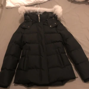 Svart dunjacka från Moose Knuckles - Selger en stilig svart dunjakke fra Moose Knuckles med en myk pelsdetalj på hetten. Jakken har glidelås foran og er perfekt for kalde vinterdager. Den er i veldig god stand og har et stilig design med logo på ermet. Kun brukt 2/3 ganger helt ny jakke uten skader 