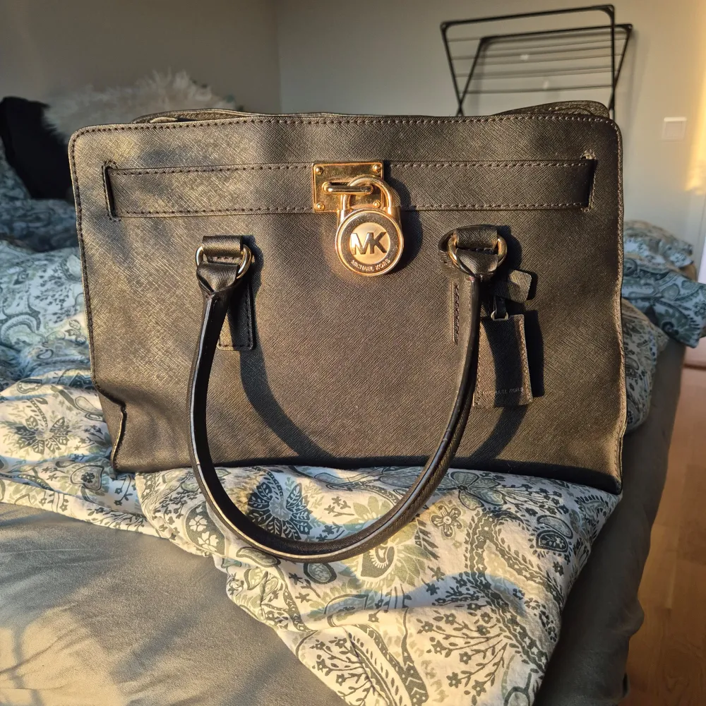 Snygg svart handväska från Michael Kors. Väskan har ett elegant lås med märkets logotyp i guld och praktiska handtag. Insidan är rymlig med flera fack och dragkedja för säker förvaring. Perfekt för både vardag och fest!. Väskor.