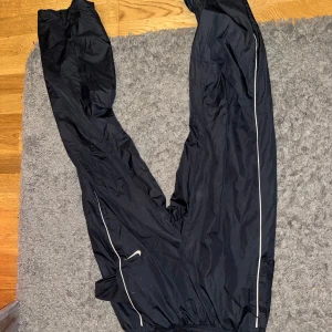 Svarta Nike trackpants  - Säljer ett par svarta träningsbyxor i syntetmaterial. De har en praktisk dragkedja längs benen och elastisk midja för bekväm passform. Perfekta för utomhusträning eller löpning. Ändå problemet är att det finns ett litet hål mellan benet men det sys ihop vid beställning