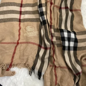 Beige rutig halsduk  Burberry - Säljer en klassisk beige rutig halsduk. Den har det ikoniska rutmönstret i svart, röd och vit. Perfekt för att hålla sig varm och stilren under kyliga dagar. Halsduken är i bra skick och passar perfekt till höst- och vintergarderoben.