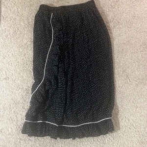 Svart prickig kjol med volang - Säljer en superfin svart kjol med vita prickar och volangdetaljer. Kjolen har en söt vit kant som ger den lite extra stil. Perfekt för både vardag och fest! 🖤funkar även att ha som klänning för det är resår i midjan!!