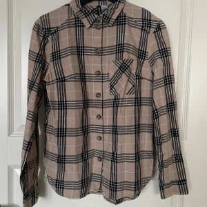 Beige och svart rutig skjorta från Divided - Säljer en snygg beige och svart rutig skjorta från Divided. Skjortan har långa ärmar och knappar framtill. Perfekt för höst och vår, och den är i mycket bra skick. Passar både till vardags och lite mer uppklädda tillfällen.