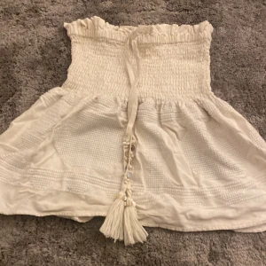 Vit kjol med tofsdetaljer - Säljer en superfin vit kjol med elastisk midja och dekorativa tofsar. Kjolen har ett snyggt texturerat mönster och små pärldetaljer som ger en bohemisk känsla. Perfekt för sommardagar eller en avslappnad strandlook. 🏖️