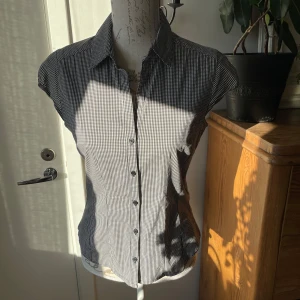 Svartvit rutig blus från H&M - Säljer en snygg svartvit rutig blus från H&M i storlek S. Blusen har korta ärmar och knappar framtill. Perfekt för både vardag och festliga tillfällen. Den är i mycket bra skick och gjord av bekvämt material. 70% bomull, 14% polyester, 13% polyamid, 3% elastan 
