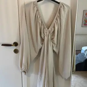 Säljer en elegant beige klänning med djup v-ringning och långa ärmar. Klänningen har en snygg knytning framtill som ger en fin detalj. Perfekt för både vardag och festliga tillfällen. Materialet känns mjukt och bekvämt mot huden.