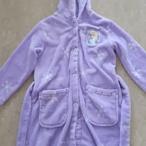 Supermysig lila fleece morgonrock från Disney med Elsa från Frost på bröstet. Den har långa ärmar och knappar framtill, samt två praktiska fickor. Perfekt för kalla morgnar eller mysiga kvällar.