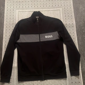 Svart tröja med dragkedja från BOSS - Säljer en stilren svart tröja från BOSS med en grå rand och logga över bröstet. Tröjan har en dragkedja framtill och långa ärmar, perfekt för både vardag och fest. Den är i mycket bra skick och passar till alla tillfällen.