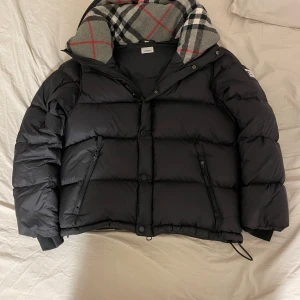 Svart pufferjacka från Burberry - Säljer en snygg svart pufferjacka från Burberry i mycket bra skick. Jackan har avtagbara ärmar, vilket gör den till en väst också. Den har en dragkedja och knappar framtill, samt en huva med ett klassiskt Burberry-rutigt mönster. Perfekt för kalla dagar! 🖤 pris kan diskuteras!!! 