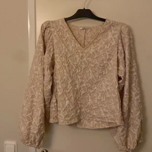 Beige blus med blommigt mönster - Säljer en superfin beige blus med ett diskret blommigt mönster. Den har långa ärmar och en v-ringad hals. Perfekt för både vardag och fest! 🌸