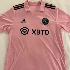 Säljer en snygg rosa fotbollströja från Adidas med korta ärmar. Den har svarta detaljer som tre ränder på axlarna och logotyper på bröstet. Perfekt för fotbollsfans som vill sticka ut på planen eller läktaren. Tröjan är i mycket bra skick och redo för nya äventyr!