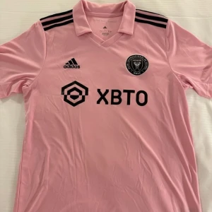 Rosa fotbollströja från Adidas - Säljer en snygg rosa fotbollströja från Adidas med korta ärmar. Den har svarta detaljer som tre ränder på axlarna och logotyper på bröstet. Perfekt för fotbollsfans som vill sticka ut på planen eller läktaren. Tröjan är i mycket bra skick och redo för nya äventyr!