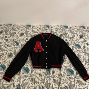 Svart bomberjacka med röd detalj - Snygg svart bomberjacka med röd randig detalj på krage, ärmslut och nederkant. Har en stor röd bokstav 'A' på bröstet och knappar framtill. Perfekt för en cool och avslappnad stil!