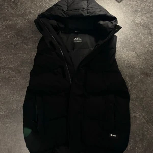 Svart dunväst från Zara - Säljer en svart dunväst från Zara i mycket bra skick. Västen har en dragkedja och en mysig huva, perfekt för höst och vinter. Den är stilren och passar till de flesta outfits. Perfekt för lager-på-lager-stil!