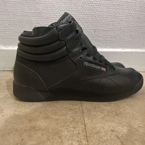 Svarta Reebok Classic sneakers - Säljer ett par svarta Reebok Classic sneakers i bra skick. De har en hög modell med snörning och kardborreband för extra stöd. Perfekta för både vardag och träning. Klassisk design med Reebok-logga på sidan och tungan.