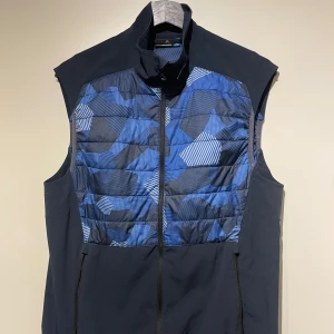 Blå mönstrad väst från J.Lindeberg - Blå golfväst från J.Lindeberg med ett coolt  mönster i olika blå nyanser. Västen har en dragkedja framtill. Perfekt för lager-på-lager under alla fyra årstider. Märkeslogga synlig på både fram- och baksida. Passar både till vardags, träning å framförallt golf!