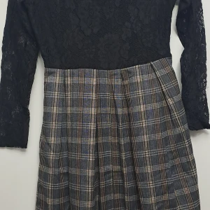 Svart spetsklänning med rutig kjol - Snygg klänning med svart spets upptill och långärmad design. Kjolen är rutig i grå och blå toner, vilket ger en klassisk look. Perfekt för höst och vinter, kanske till en fest eller julfirande. Klänningen har en normal passform och är både elegant och bekväm.
