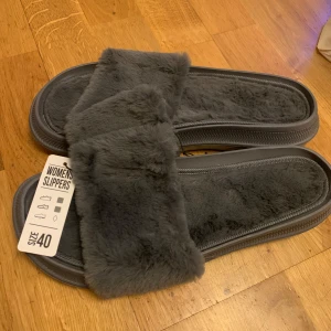 Grå damtofflor i syntetpäls - Mysiga grå damtofflor i storlek 40 med mjuk syntetpäls. Perfekta för att hålla fötterna varma och bekväma hemma. De har en stabil sula och en bred rem över foten för extra komfort. Perfekta för kalla dagar inomhus!