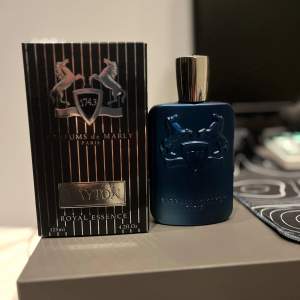 Säljer en lyxig parfym från Parfums de Marly, modellen Layton. Flaskan är elegant i en djupblå färg med en stilren design och ett silverfärgat lock. Förpackningen är svart med silverdetaljer och har en kapacitet på 125 ml. Perfekt för den som vill ha en sofistikerad doft med en touch av elegans.