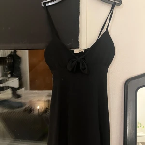 Svart klänning med rosett - Säljer en elegant svart klänning med smala axelband och en söt rosett framtill. Klänningen har en smockad rygg för bättre passform och är perfekt för festliga tillfällen. Köpt i Italien. Aldrig använt  
