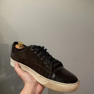 Bruna sneakers från Lanvin - Säljer ett par snygga svarta sneakers från Lanvin i mycket bra skick. Skorna har en stilren design med snörning och en vit sula som ger en cool kontrast. Perfekta för både vardag och fest! Kommer med originalpåse.