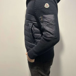 Svart Monclear cardigan  - Säljer en HELT NY och  snygg Monclear cardigan. Den har en cool kombination av stickade ärmar och perfekt för höst och vinter. Jackan har en dragkedja framtill och en praktisk ficka med dragkedja på ärmen. Den är i mycket bra skick och passar perfekt för både vardag och fest.