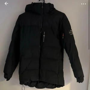 Svart dunjacka från sail racing  - Säljer en svart dunjacka från sail racing  i mycket bra skick. Jackan har en dragkedja framtill och är långärmad med justerbar huva. Perfekt för kalla vinterdagar och håller dig varm och bekväm. Passar både till vardags och utomhusaktiviteter.