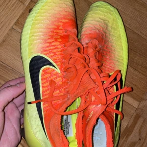 Nike fotbollsskor i orange och gul - Säljer ett par Nike fotbollsskor i en cool orange och gul färgkombination. Skorna har snörning och är perfekta för fotbollsträning eller match. De har en snygg svart Nike-logga på sidan och är i bra skick. Perfekta för dig som vill sticka ut på planen! Storlek 42 och sällsynt modell av Nike magista som inte säljs längre.