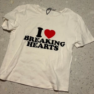 Vit t-shirt från Stradivarius - Säljer en vit t-shirt från Stradivarius med texten 'I ❤️ BREAKING HEARTS' i svart och rött. Den är kortärmad och gjord i bomull, perfekt för en avslappnad stil. Passar bra till både jeans och kjol!