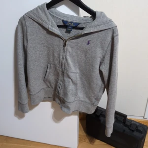 Grå hoodie från Ralph Lauren - Säljer en snygg grå hoodie från Ralph Lauren i storlek L men är mycket mindre passar s/m. Den har en dragkedja framtill och en klassisk broderad logga i lila på bröstet. Perfekt för höst och vår, och den är i mycket bra skick. Superbekväm och stilren, perfekt för vardagsbruk!