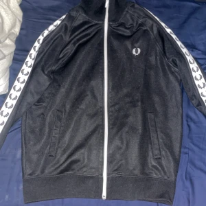 Svart track jacket från Fred Perry storlek M - Säljer en svart track jacket från Fred Perry i mycket bra skick. Den har vita detaljer längs ärmarna med det klassiska Fred Perry-mönstret. Jackan har dragkedja framtill och är perfekt för både träning och vardag. Storleken är herr M och den är riktigt bekväm och stilren.