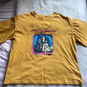 Säljer en cool gul t-shirt med Aerosmith-tryck på framsidan. T-shirten har en vintage look med ett färgglatt motiv av bandet i en TV-ruta. Perfekt för dig som älskar rock och retrostil! Passar bra till både vardags och konsert.