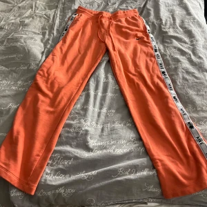 Orange mjukisbyxor från Nike - Säljer ett par orange mjukisbyxor från Nike. De har en vit och svart rand med Nike-loggan längs sidorna. Byxorna är loose fit och har en dragsko i midjan. Perfekta för chill dagar hemma eller lätt träning. Det finns några fläckar på baksidan som säkert gå att få bort, men de är fortfarande bekväma och snygga.