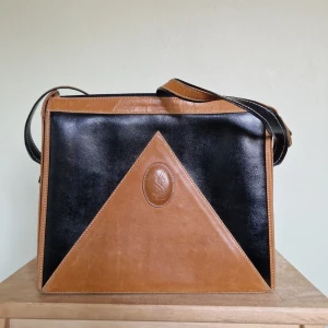 Vintage -Svart och brun axelväska med dragkedja (YSL - Osäker på äkthet!) - Snygg axelväska i svart och brunt skinn med en unik triangeldesign på framsidan. Väskan har en praktisk dragkedja upptill och en justerbar axelrem. Perfekt för både vardag och speciella tillfällen! Den hittades på Second Hand och jag vet inte om den är äkta eller inte. Värderare har varit oense om äktheten. Se min andra annons på tillhörande plånbok!