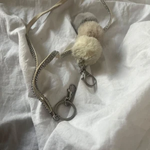 Nyckelring med fluffiga bollar och strass - Superfina nyckelringar en med två fluffiga bollar i beige och vit. Den andra har en rem dekorerad med glittriga strassdetaljer och i silverfärg. Perfekt accessoar för att piffa upp väskan eller nyckelknippan!