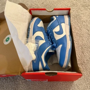 Nike Dunk Low University Blue - Säljer ett par Nike Dunk låga sneakers i en snygg blå och vit färgkombination. Skorna är i nytt skick och kommer med originalkartong. Perfekta för både vardag och festliga tillfällen. Skostorlek är herr 42.5. Passa på att köpa dessa stilrena sneakers!