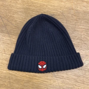 Svart ribbad mössa med Spiderman-märke - Säljer en svart ribbad mössa med ett coolt Spiderman-märke framtill. Perfekt för att hålla värmen under höst och vinter. Mössan är i bra skick och passar de flesta tack vare sin stretchiga passform.