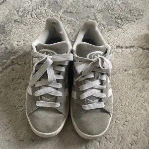 Grå sneakers från Adidas - Säljer ett par grå Adidas campus 00 då de inte kommer till användning. Köpta för ungefär 1 halvår sen och har inga synliga defekter på utsidan men har hål på insidan av hälen därav priset. Man får med gråa snören till skorna också! 