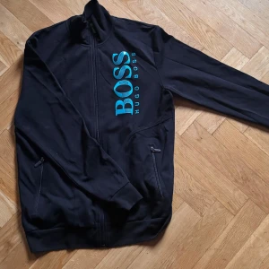 Svart tröja från Hugo boss, hel zip - Säljer en svart Hugo Boss jacka i mycket bra skick. Den har en dragkedja framtill och stora blå bokstäver med märkesnamnet på bröstet. Jackan är långärmad och perfekt för höst och vår. Den är gjord i ett mjukt och bekvämt matrial. Har tröttnat på den.